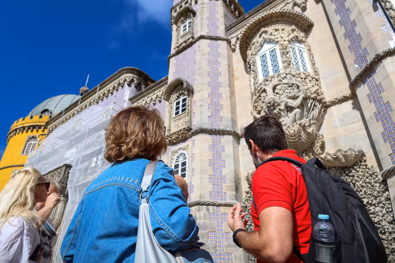 Tourists admire the intricate architecture of Pena Palace in Sintra under a clear blue sky, highlighting its ornate details.