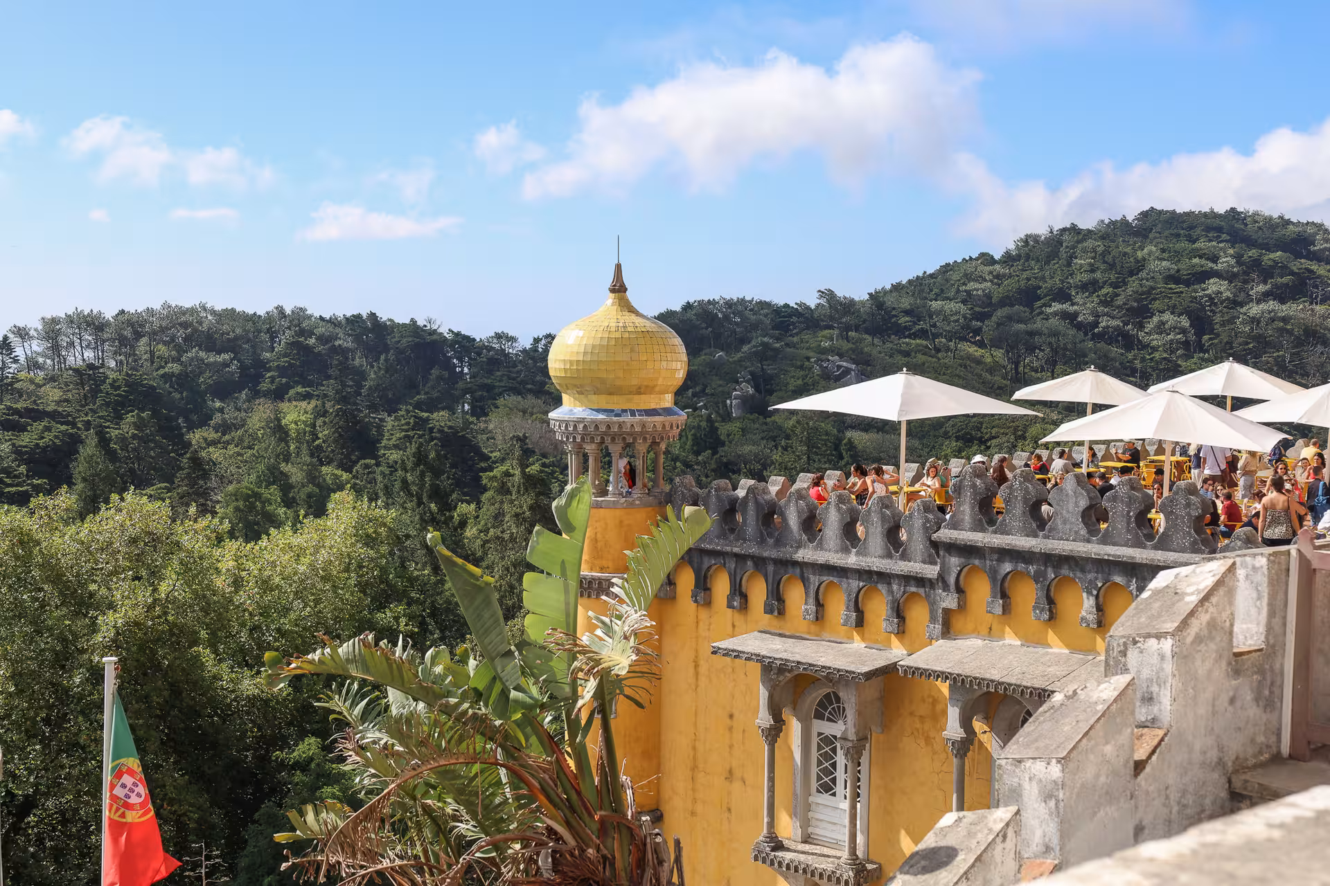 Visitors enjoy the vibrant yellow architecture and lush views at Pena Palace in Sintra, Portugal under a clear sky.