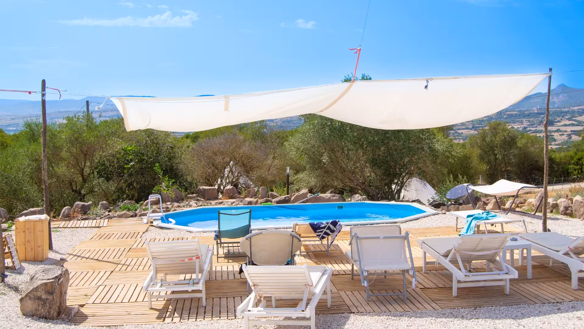 Outdoor pool area with sun loungers and shade sail at Perfugas glamping site, surrounded by scenic nature.