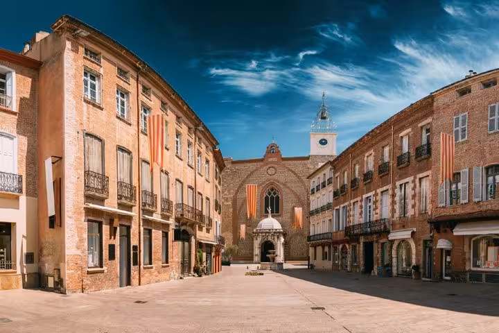 Perpignan town square with historic brick buildings, Catalan flags, and cathedral facade under blue sky