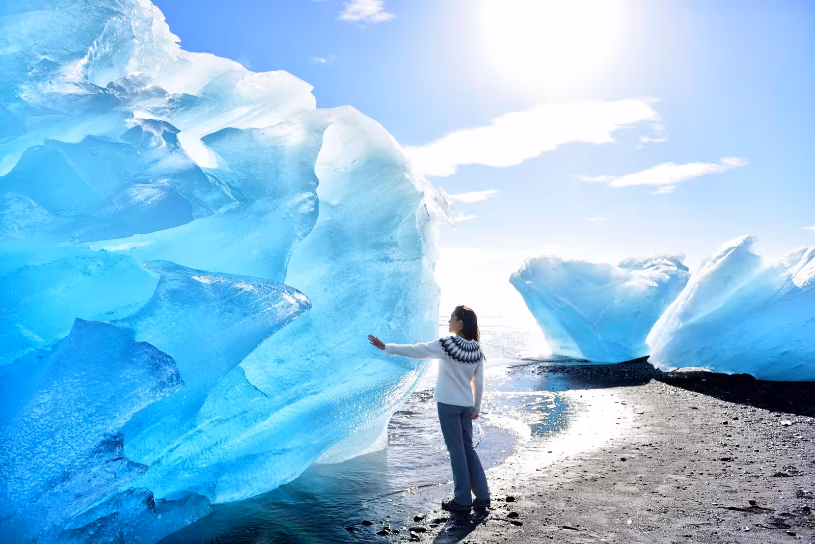 Person in cozy sweater touches massive ice formation under sunny skies on Glacier Lagoon tour.