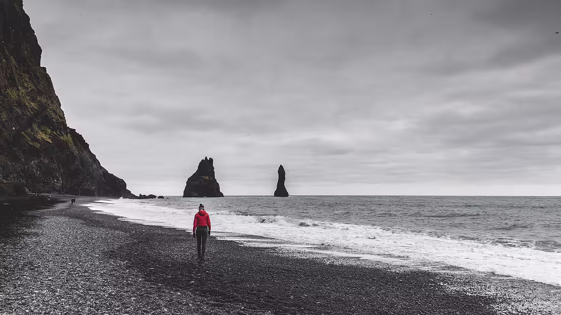 A lone person walking along a black sand beach with grey weather in the background