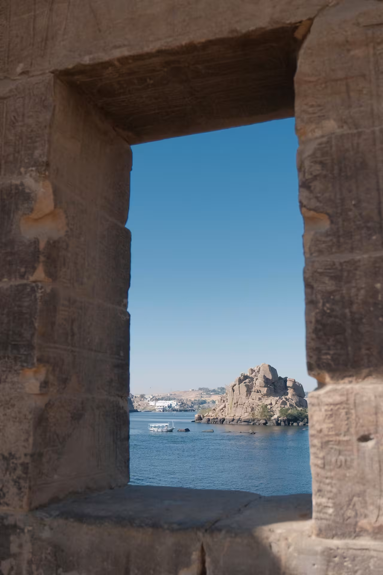 View through ancient stone window at Philae Temple overlooking the Nile River and rocky island in Aswan