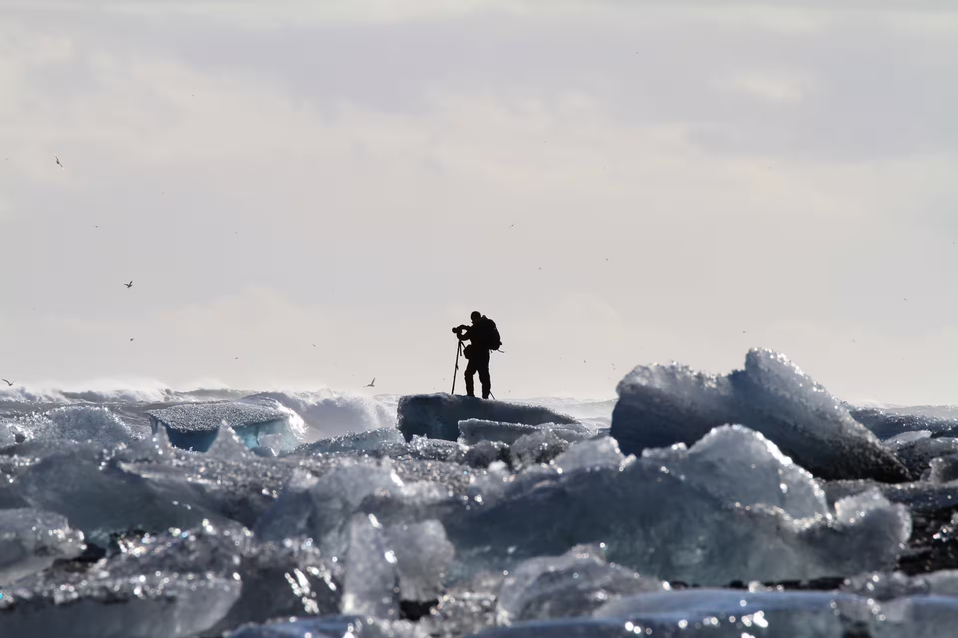 Silhouetted photographer on ice chunks at Iceland's glacier lagoon with crashing waves behind