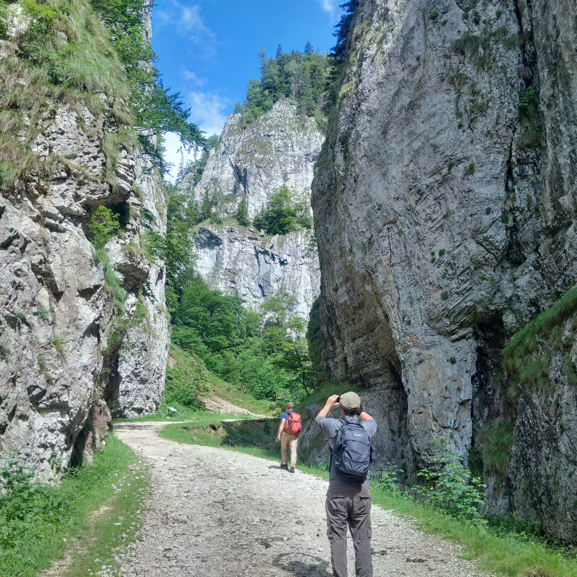 Two hikers on a rocky gorge trail surrounded by tall limestone cliffs and green trees in Piatra Craiului
