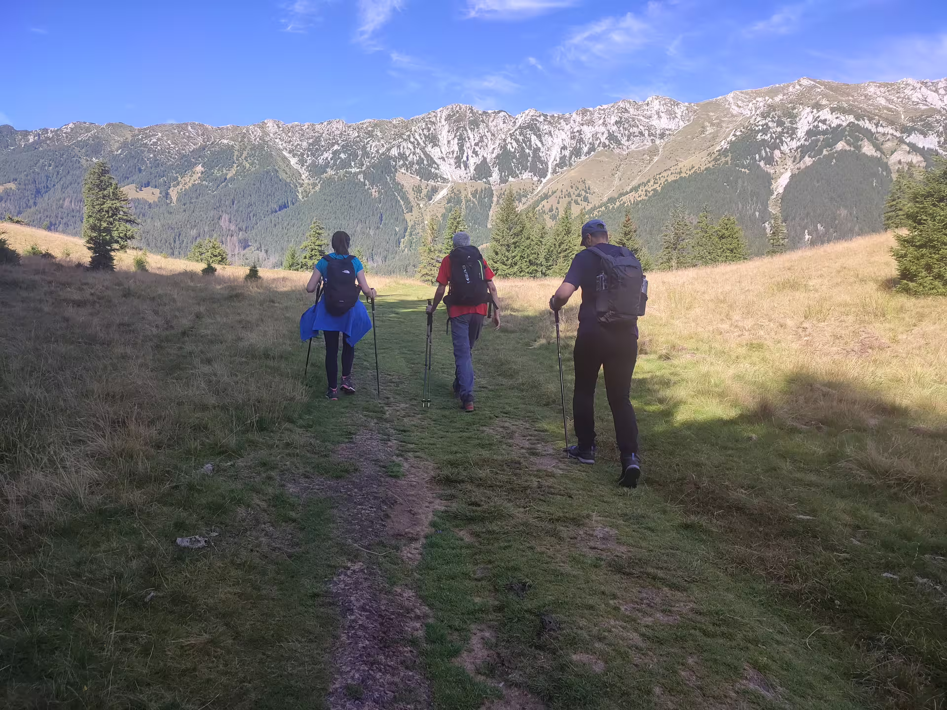 Three hikers with trekking poles and backpacks walking toward Piatra Craiului mountain ridge on a sunny day