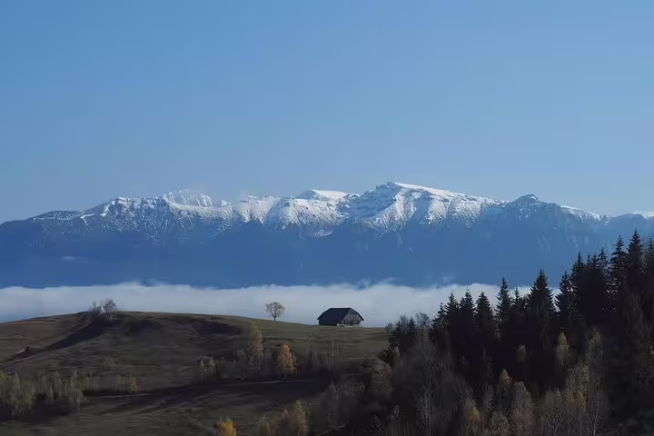 Snow-capped Piatra Craiului mountains above cloud layer with lone cabin on green hillside near Brasov