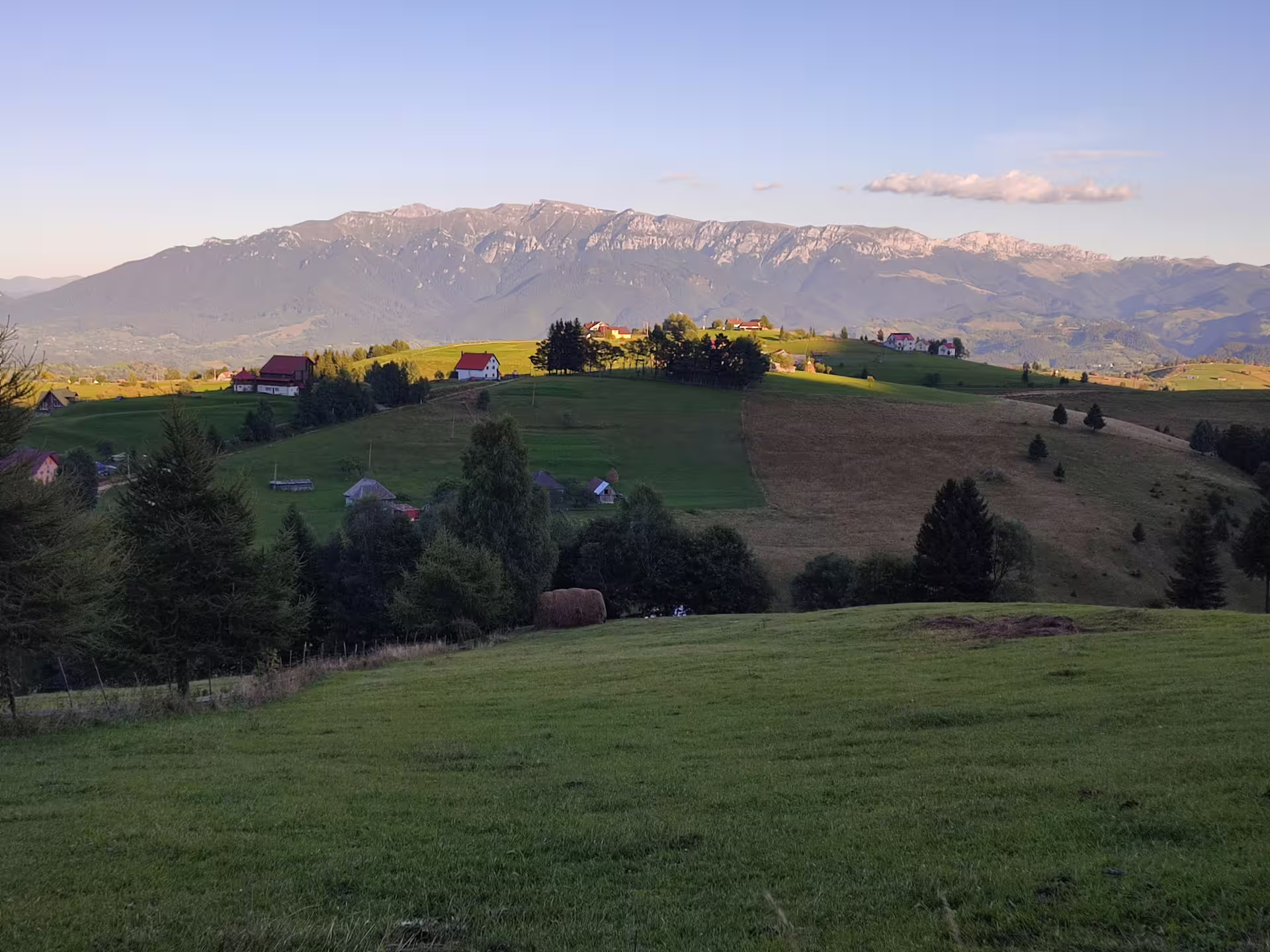 Green rolling hills with farmhouses and Piatra Craiului rocky mountain ridge backdrop near Brasov, Romania