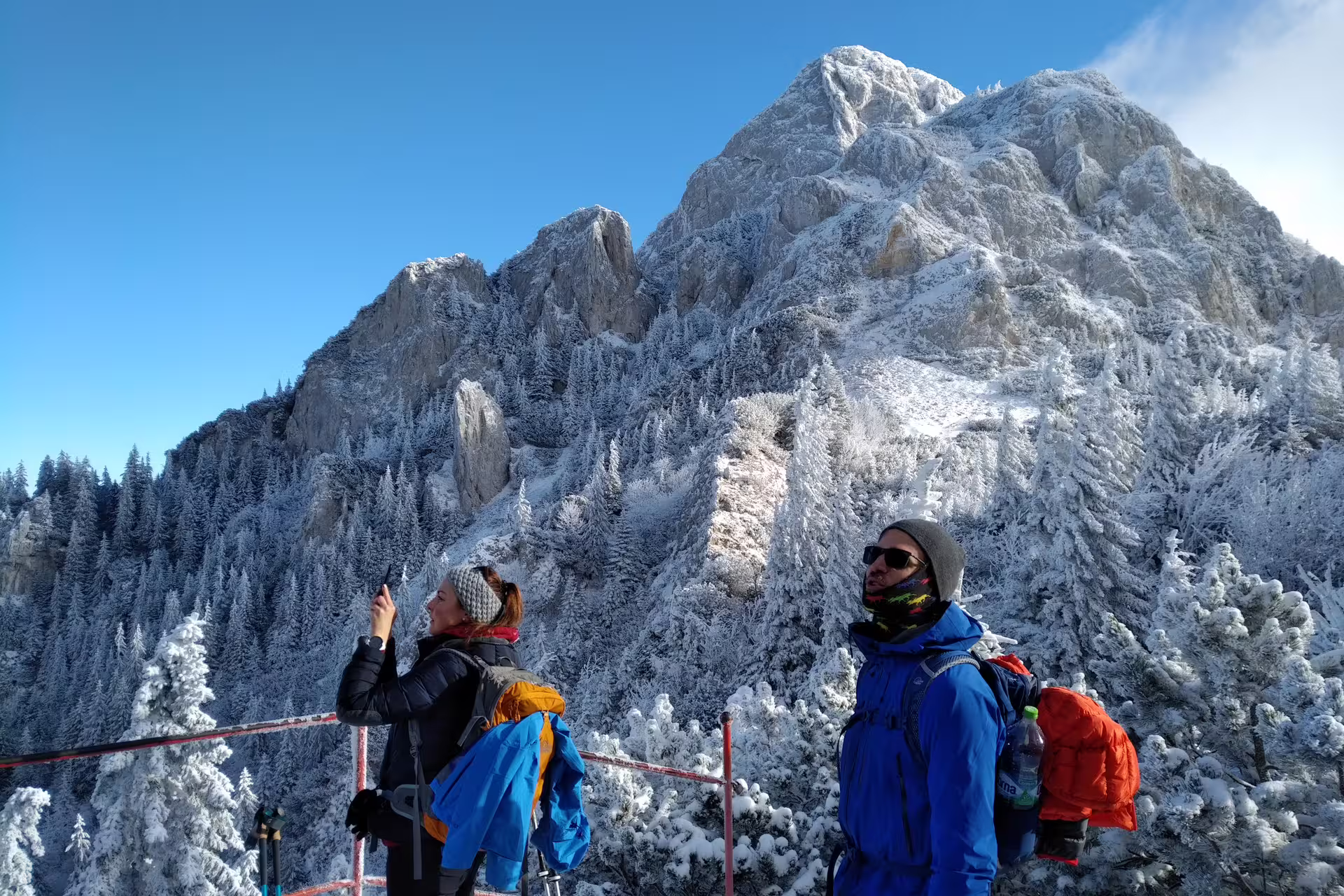 Two hikers in winter gear photographing snow-covered Piatra Craiului mountain peak on a guided trek