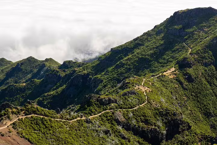 A winding path stretches across green mountain ridges at Pico do Arieiro, surrounded by clouds and blue sky.