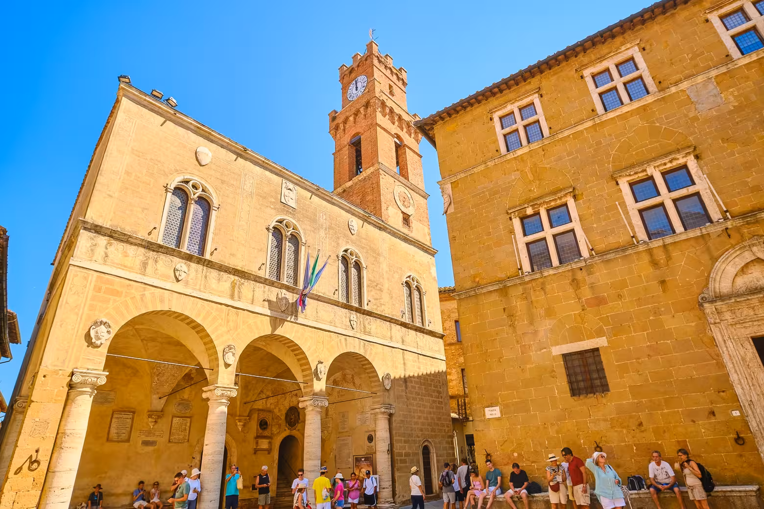 Tourists explore the historic Piazza Pio II in Pienza, Tuscany, with its iconic Renaissance architecture under a clear blue sky.