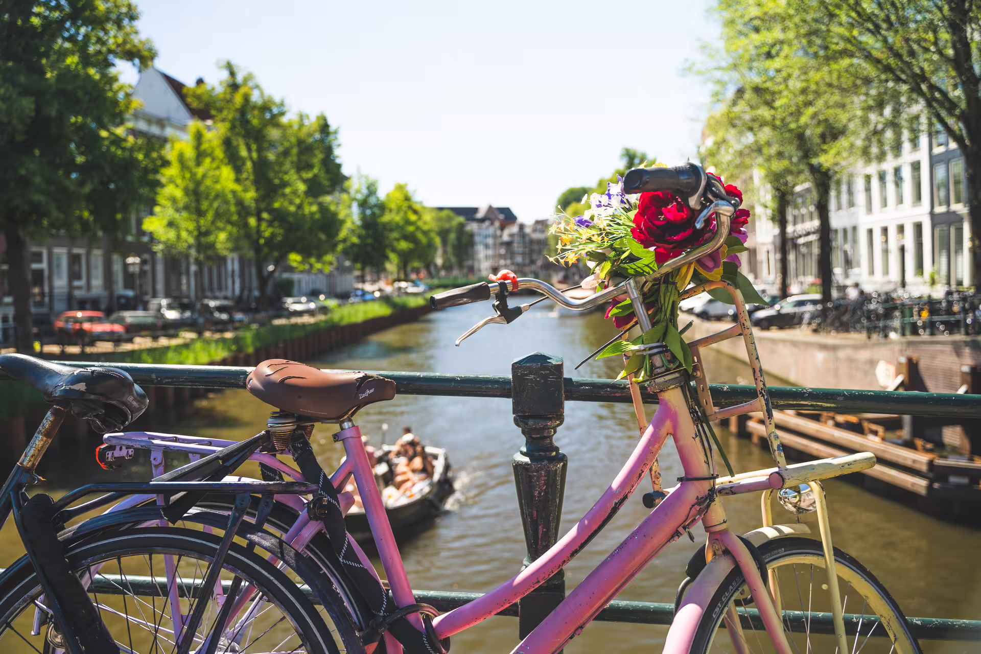 Pink bike adorned with flowers on an Amsterdam canal bridge, with a boat cruising and historic buildings in the background.
