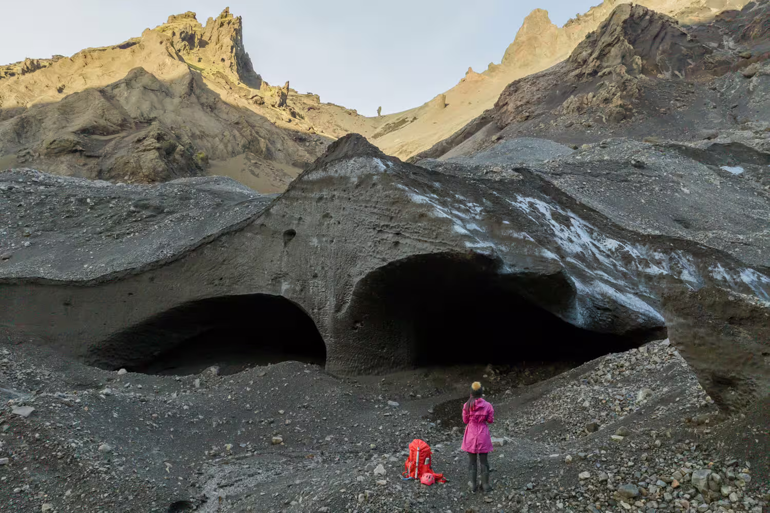 Hiker in pink jacket exploring dark ash-covered ice caves at Krossarjokull Glacier on a citizen science day trip in Iceland