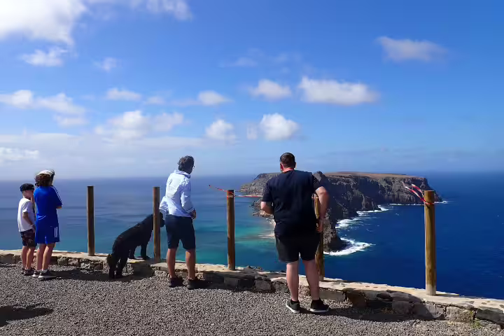 Tourists and a dog admire stunning ocean views from a cliff on the Pirate Expedition: Full Island Tour under a clear blue sky.