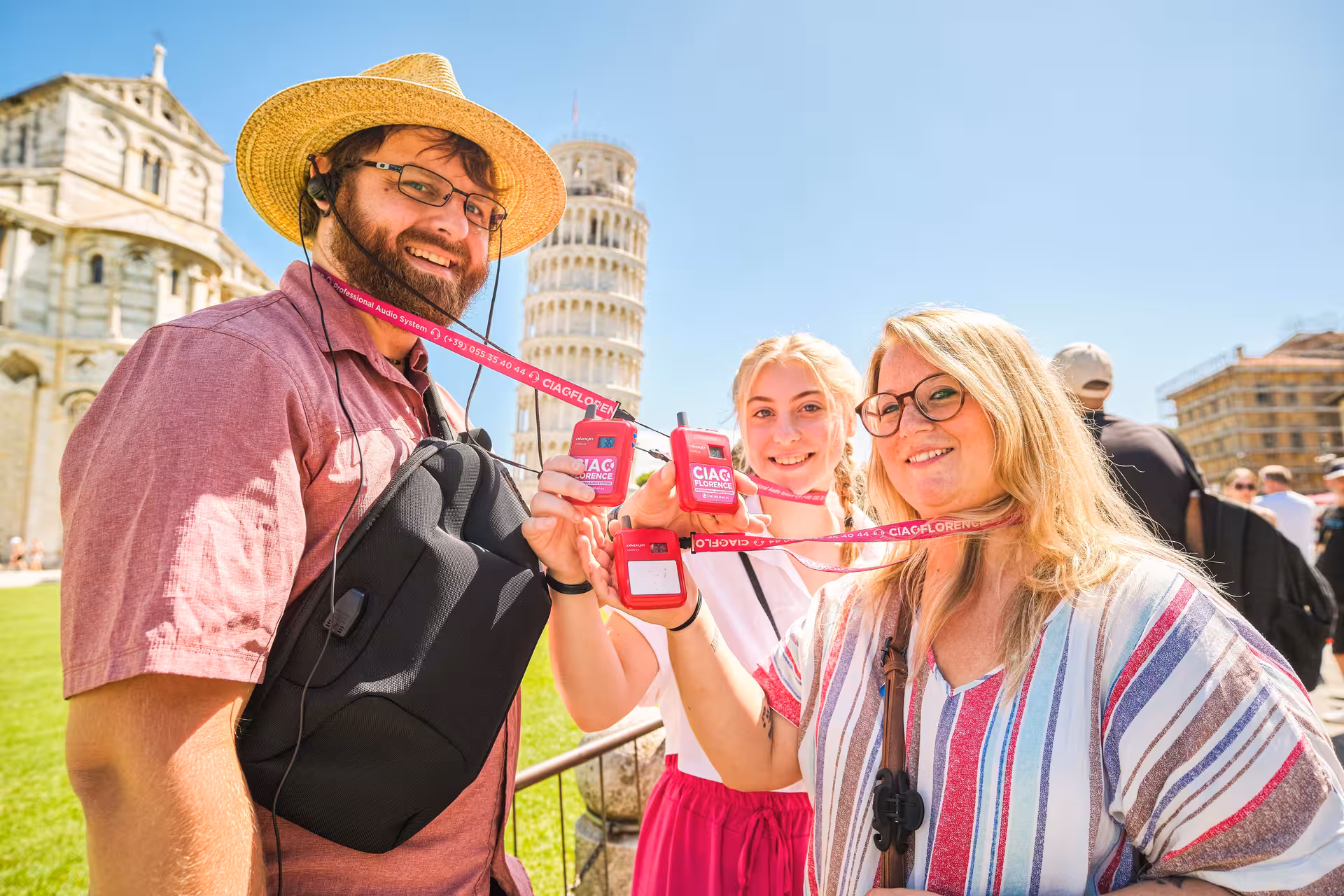 Happy tourists display tour passes in front of the iconic Leaning Tower of Pisa on a sunny day.