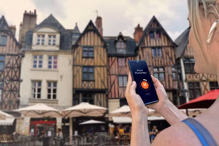 Tourist holding smartphone with audio tour app at Place Plumereau, Tours, France, with medieval half-timbered buildings