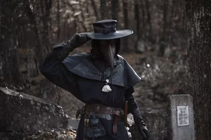 Person in black plague doctor costume with beak mask and wide-brim hat in dark forest setting