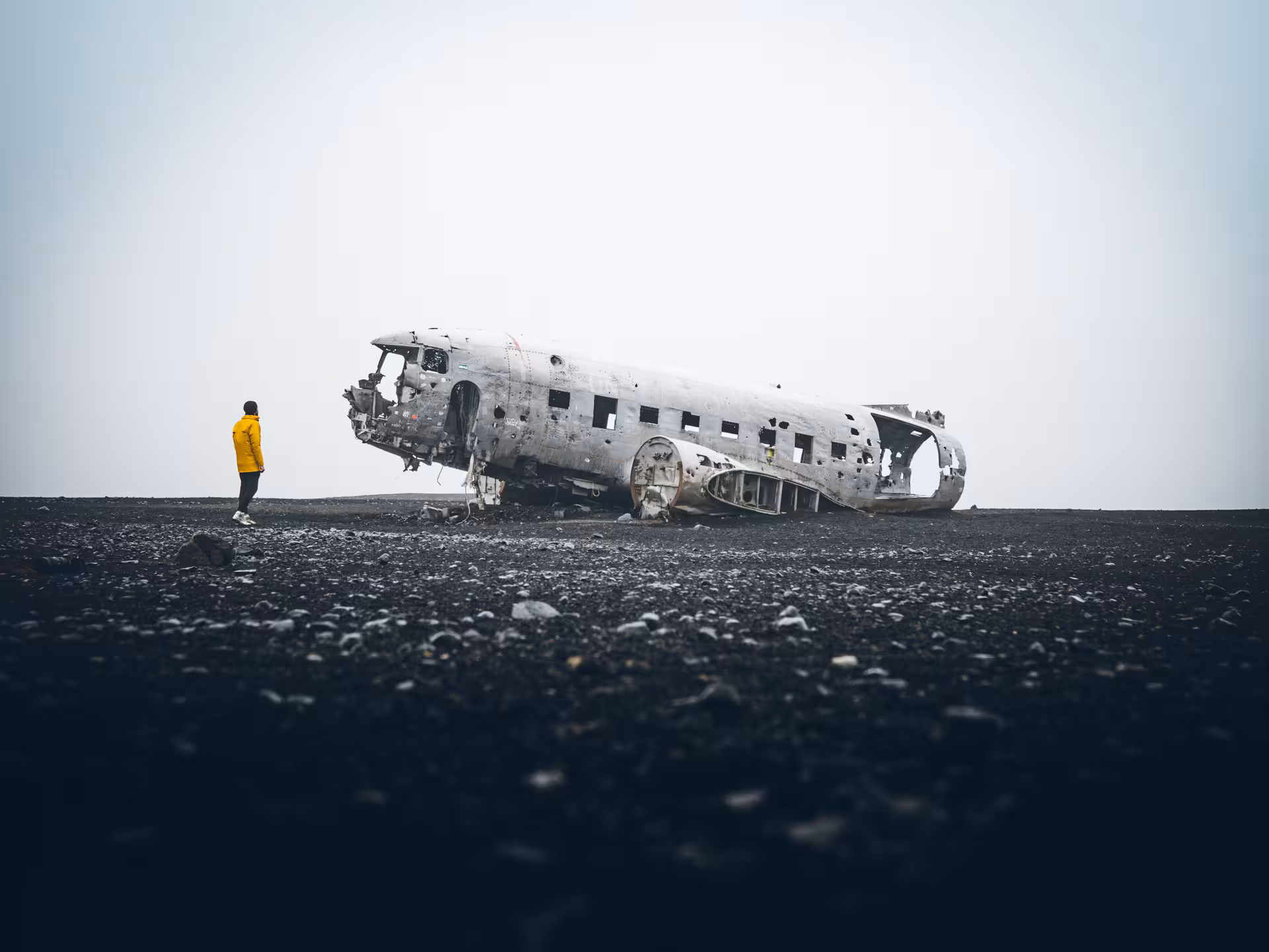 Person in yellow jacket standing beside crashed plane wreck on Iceland's black sand beach