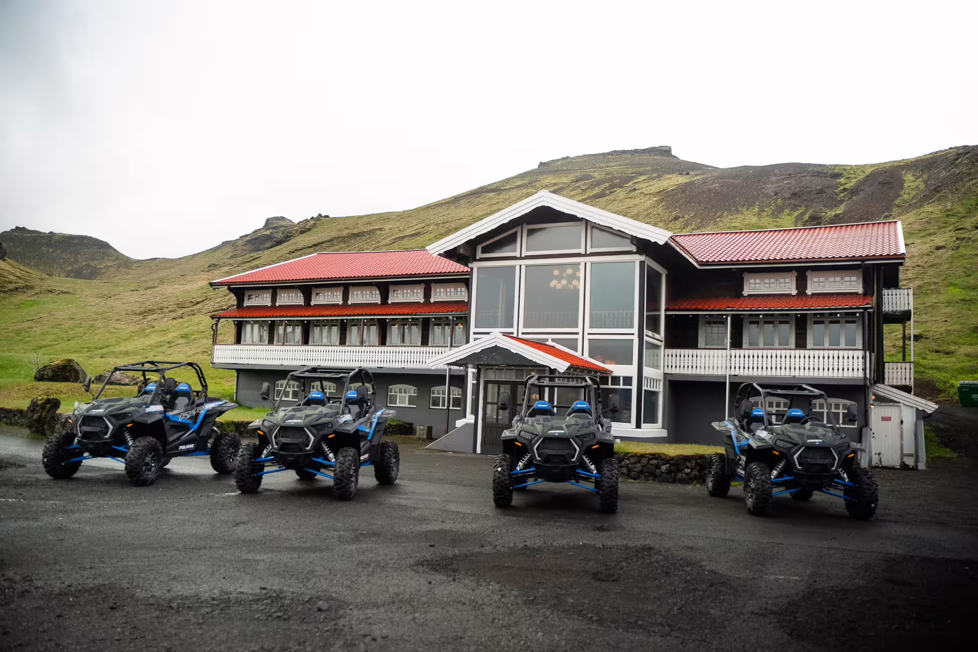 Four Polaris RZR buggy vehicles parked in front of a red-roofed lodge with green Icelandic hills behind
