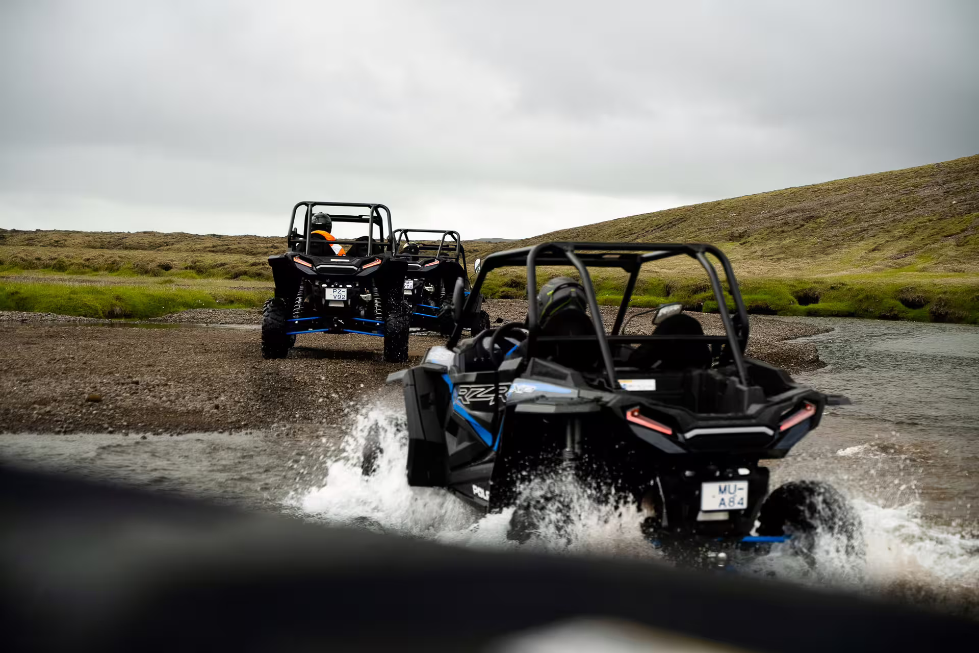 Two black Polaris RZR buggies crossing a shallow river in Iceland's rugged lava field landscape