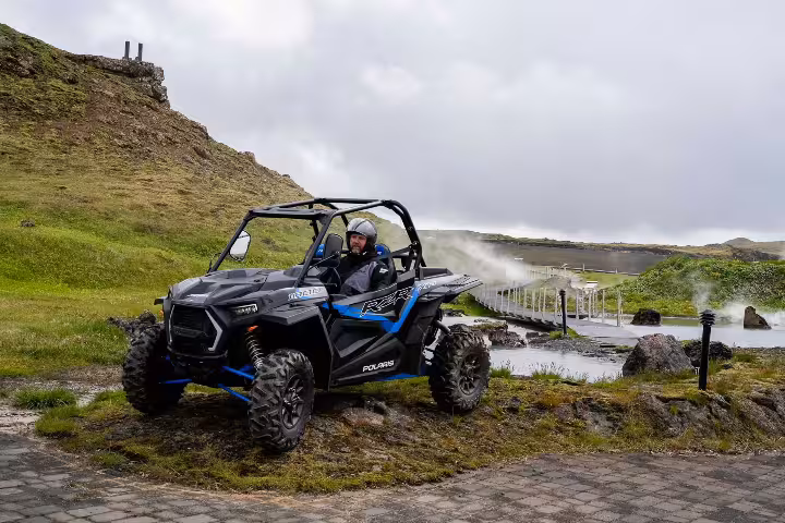 Person driving a Polaris RZR buggy on rocky terrain near a geothermal lake in Iceland