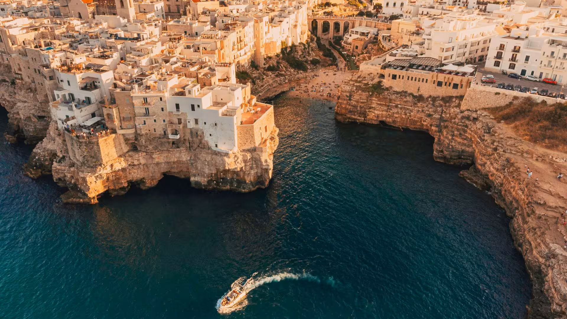 Aerial view of Polignano a Mare with a boat navigating the turquoise waters near the iconic cliffside buildings.