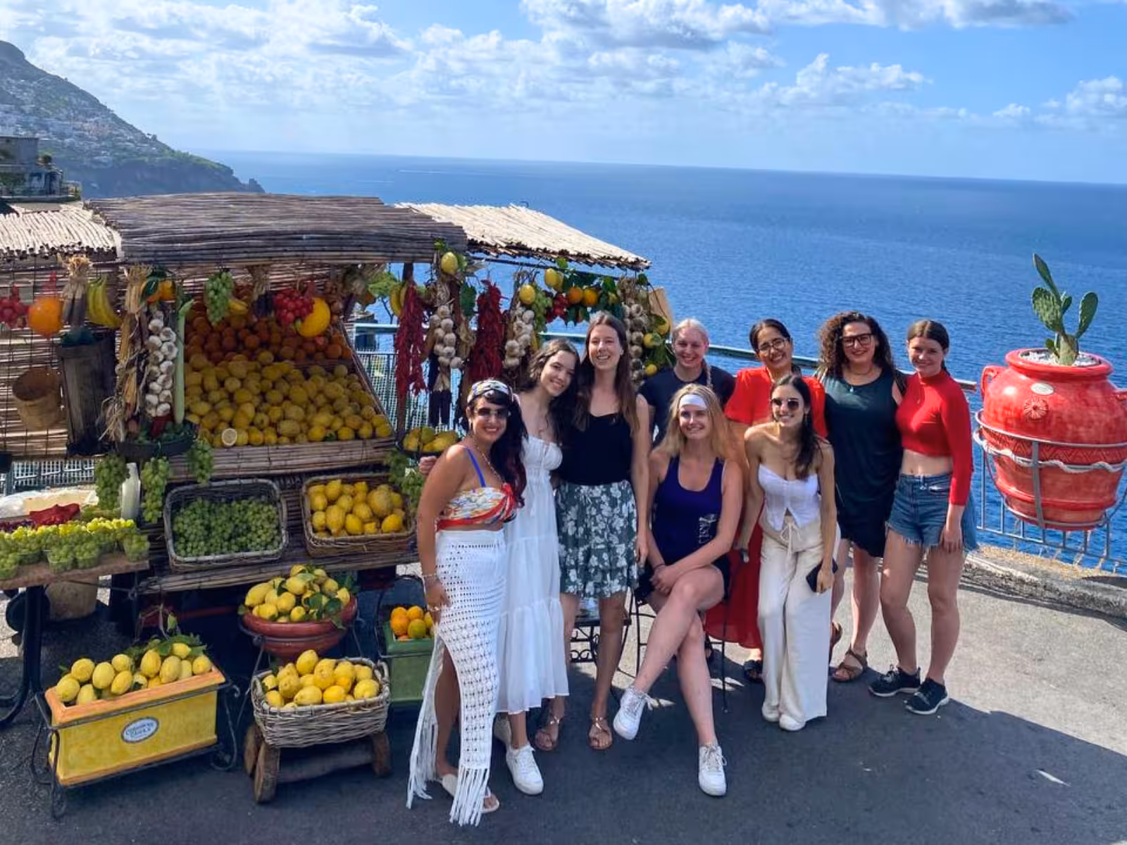 Group of tourists enjoying a vibrant fruit stand with stunning Amalfi Coast views.