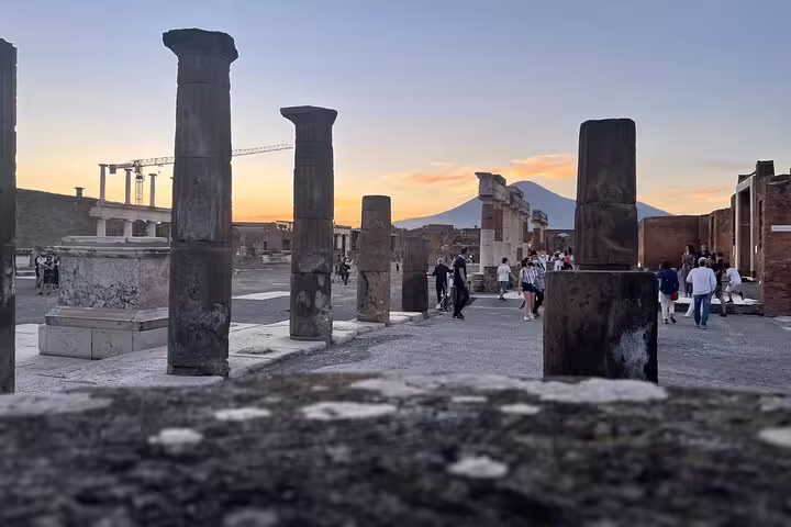 Tourists explore Pompeii's ancient columns with Mount Vesuvius silhouetted at sunset, capturing historical beauty.