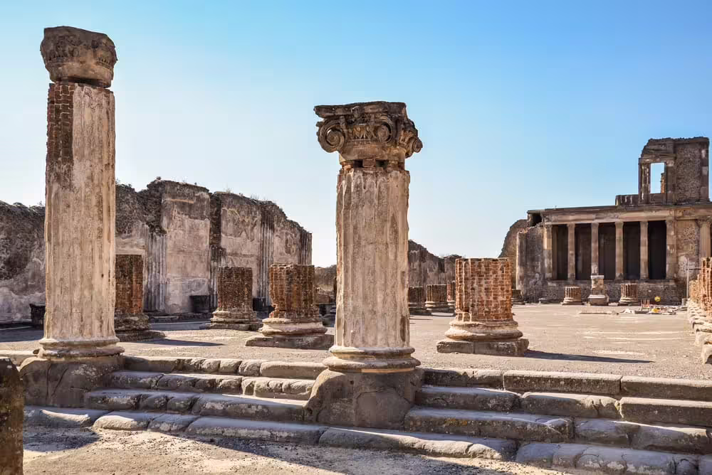 Ancient Pompeii forum with stone columns and temple ruins under blue sky, guided group walking tour in excavations