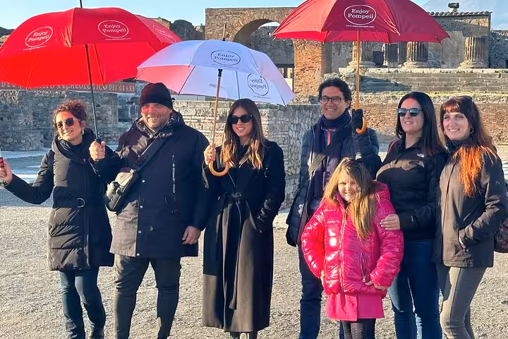 Tourists enjoy a guided tour of Pompeii, holding red and white umbrellas on a sunny day.