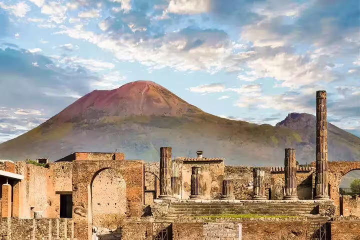 Ancient Pompeii ruins with Roman columns and arches, Mount Vesuvius volcano in background under cloudy sky