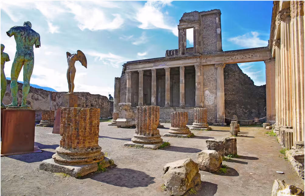 Ruins of the Temple of Apollo in Pompeii with standing statues and ancient columns under a blue sky.