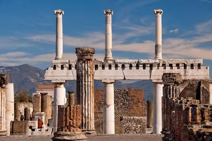 Majestic ruins of Pompeii's Temple of Jupiter with towering columns against a backdrop of mountains and blue sky.