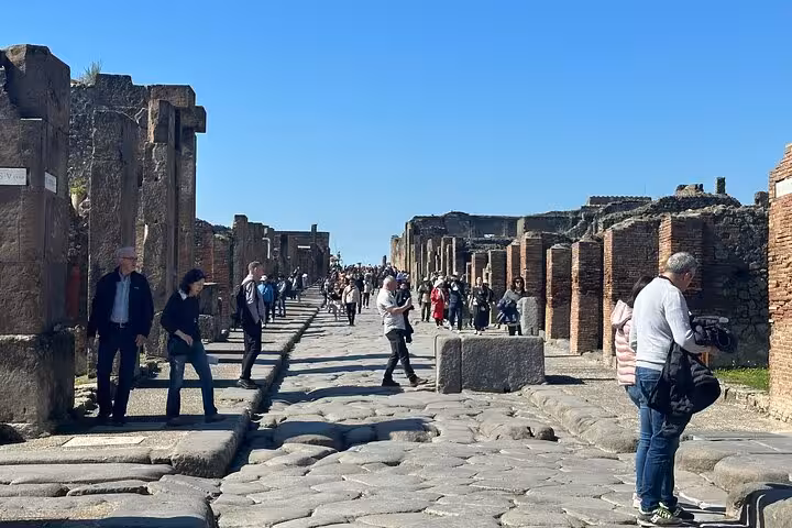 Tourists explore the ancient stone streets of Pompeii under a clear blue sky, part of the Pompeii and Vesuvius tour.