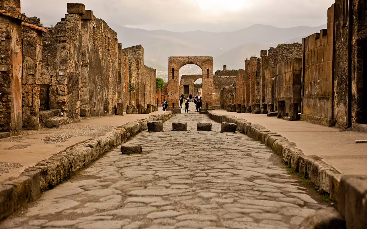 Group walking tour in Pompeii excavations along cobblestone Via dell’Abbondanza toward arch with ruins views