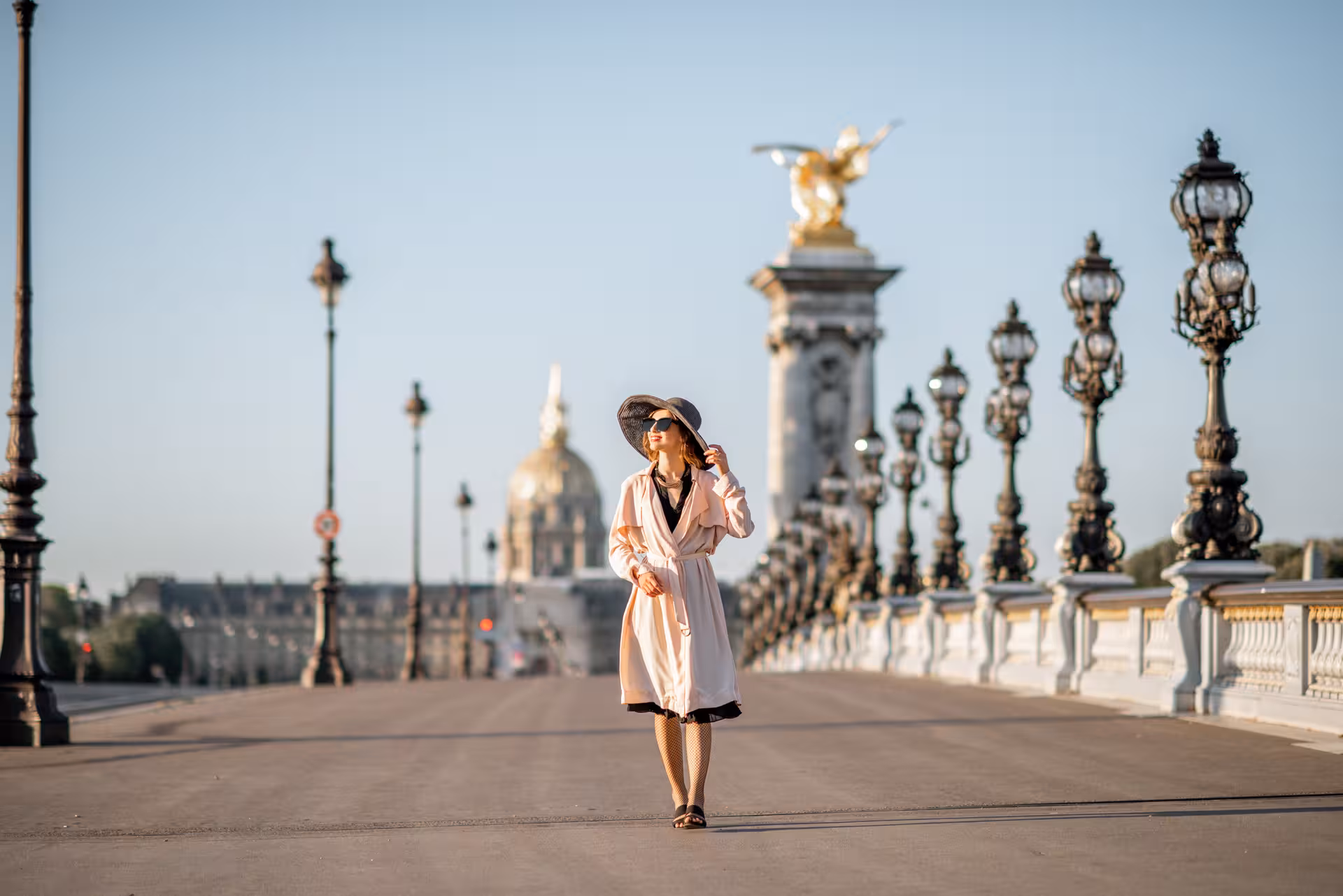 Elegant woman in pink trench coat walking on Pont Alexandre III bridge with Les Invalides golden dome in background