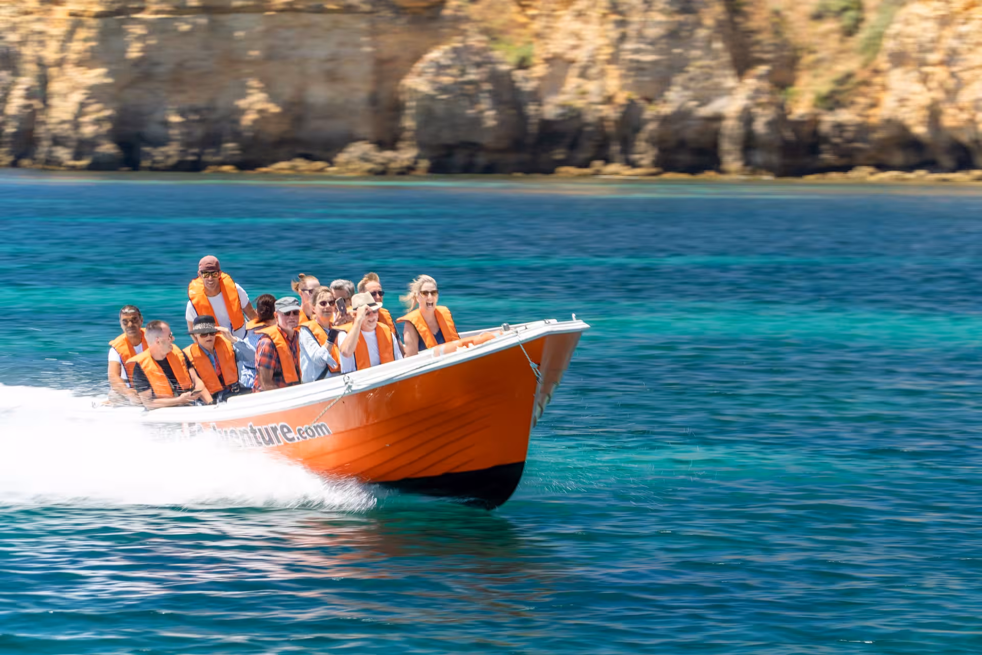 Excited tourists in life jackets speeding on an orange boat past turquoise waters and rocky cliffs on a Ponta da Piedade cave tour