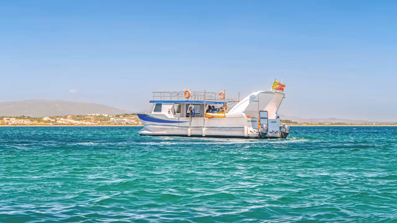 A tour support boat sailing on turquoise Lagos waters with the Portuguese coastline and clear blue sky behind.