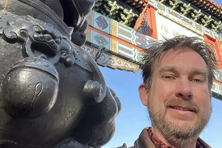 Man taking selfie beside bronze lion statue at ornate Chinese gate with colorful architecture