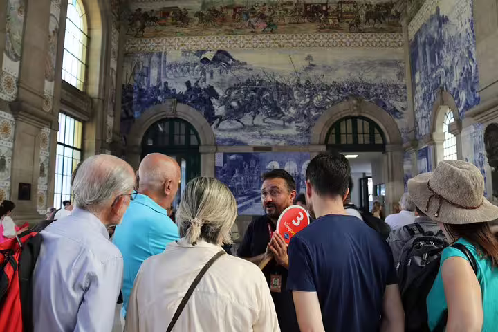 Tourists listening to a guide in a historic Porto train station adorned with stunning blue azulejo tiles.