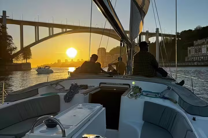 Scenic Douro River boat tour in Porto at sunset with passengers enjoying wine and snacks, passing under iconic bridge.