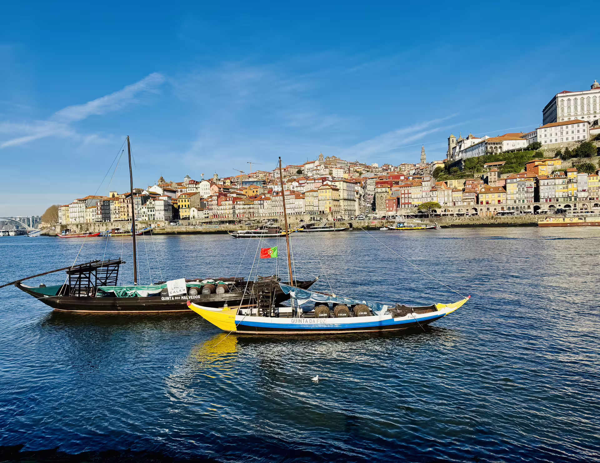 Traditional boats on the Douro River with a scenic view of Porto's colorful hillside buildings under a clear blue sky.