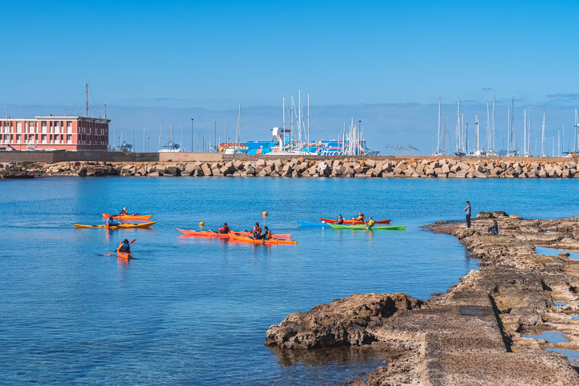 Group of kayakers navigating calm waters in Porto Torres harbor with colorful boats and scenic backdrop.