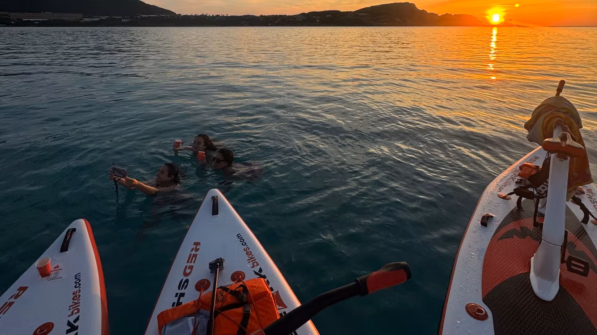 People swimming beside water bikes at sunset in Porto Torres, enjoying a scenic and relaxing experience on the water.