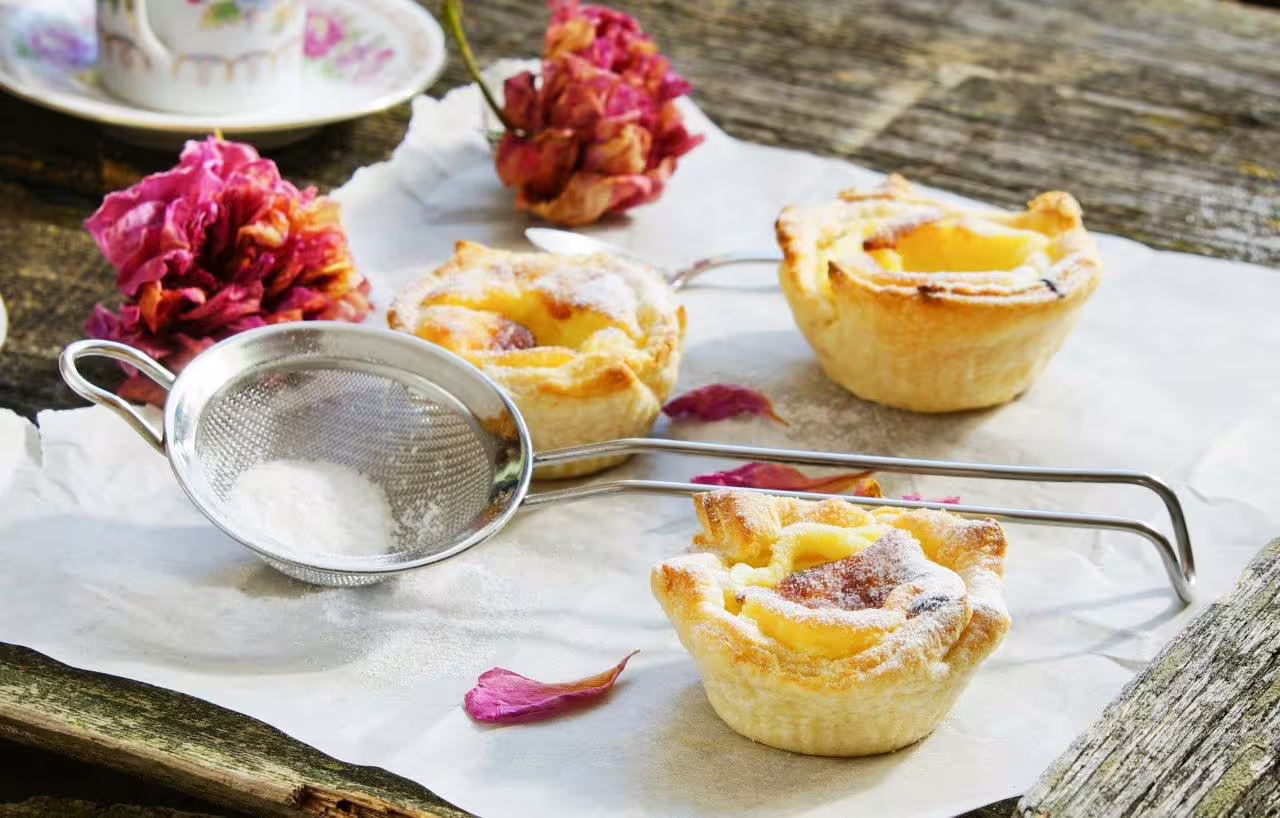 Freshly baked Portuguese pastries dusted with powdered sugar, set on rustic paper with a tea strainer and dried flowers.