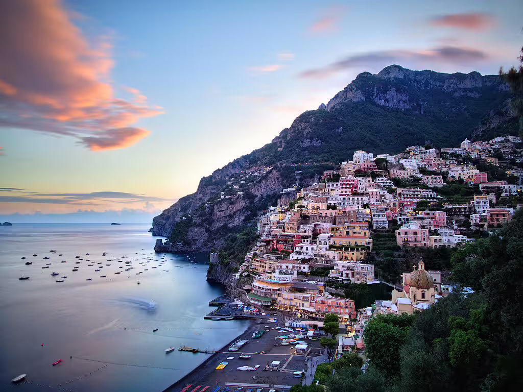 Colorful houses cascade down the cliffs of Positano at sunset, overlooking calm Amalfi Coast waters dotted with cruise boats