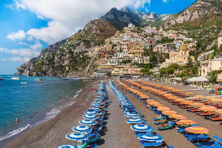 Colorful beach umbrellas on Positano's black sand beach with cliffside village and mountain backdrop, Amalfi Coast