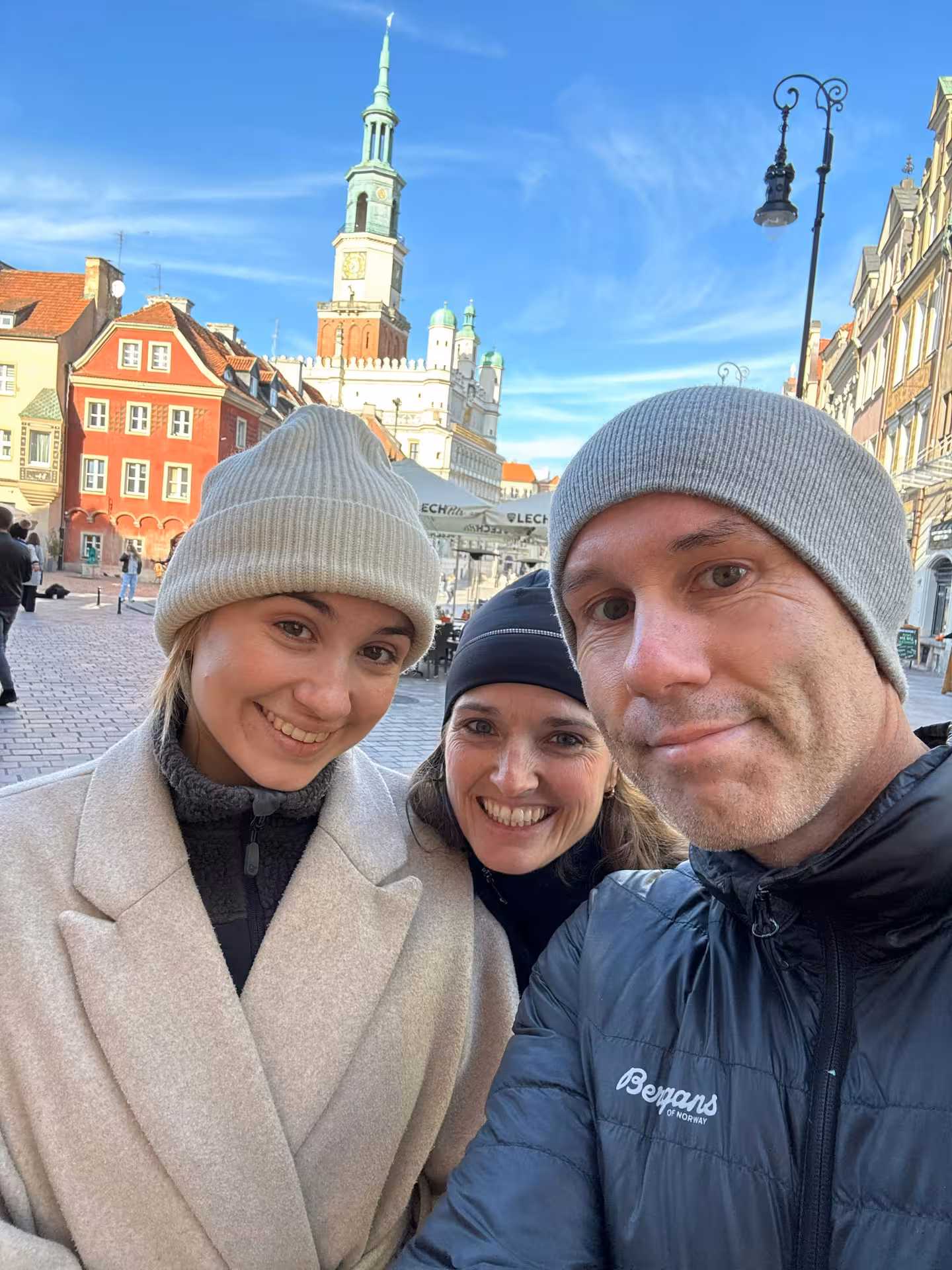 Three smiling tourists taking a selfie at Poznan Old Market Square with the historic Town Hall tower in the background