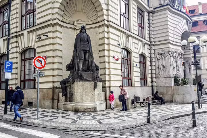 Dark bronze statue on pedestal in Prague Old Town square with cobblestone streets and historic baroque building