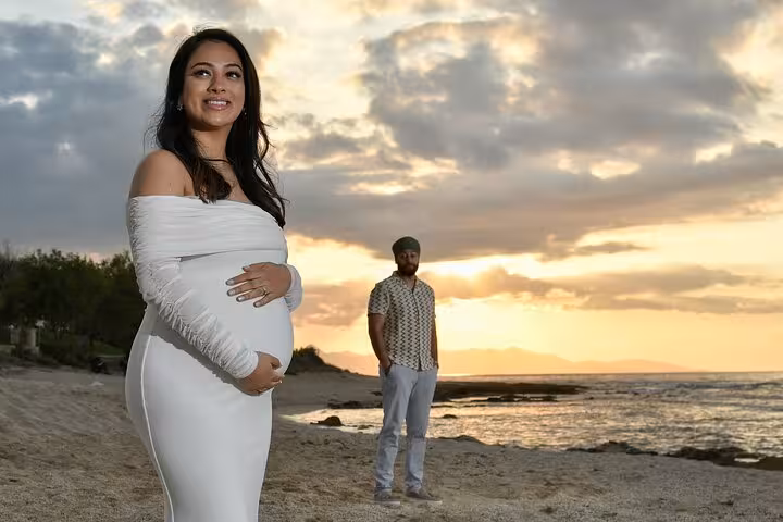 Pregnant woman smiles on Ammoudara beach at sunset, with partner in background, during a private maternity photoshoot.